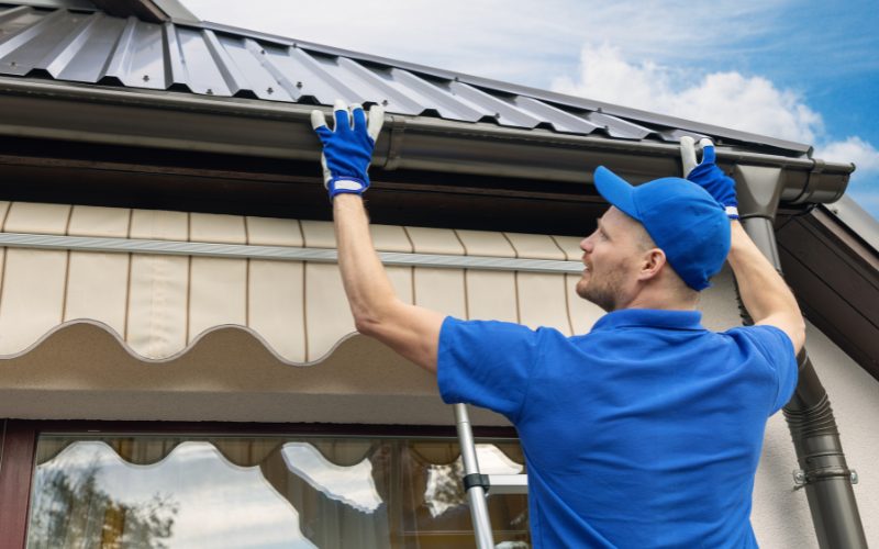 A worker in blue uniform and gloves is installing or inspecting a gutter on the edge of a roof.