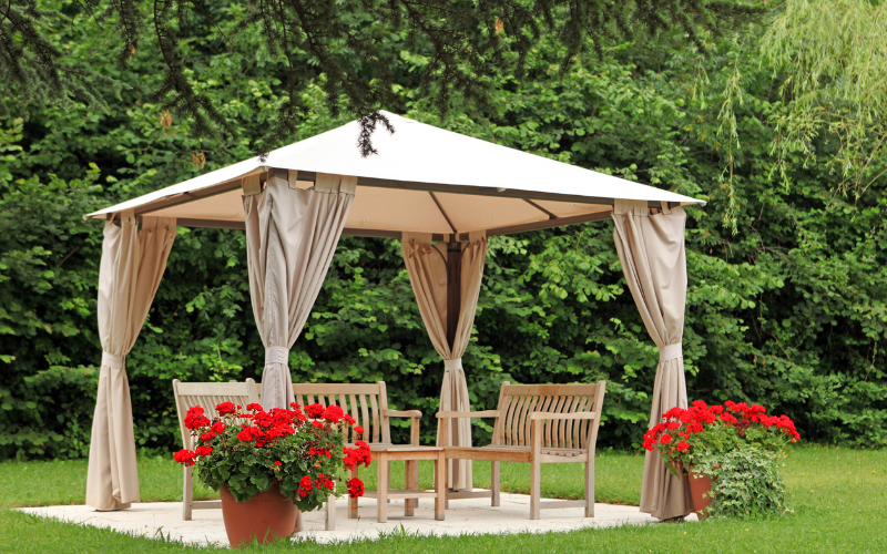 A gazebo with beige curtains is set up in a garden, surrounded by green trees and red flowers in planters.